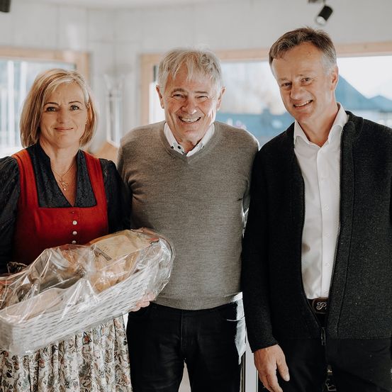 Drei Personen posieren für ein Foto. Eine Frau hält einen Korb, während zwei Männer neben ihr stehen und lächeln. Sie sind drinnen, mit Fenstern und einer Lampe im Hintergrund.