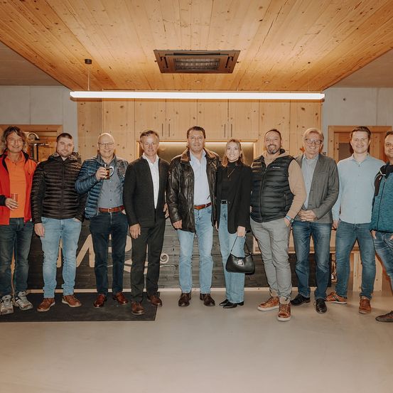 Eine Gruppe von Menschen in einem Raum, einige mit Getränken, posiert für ein Foto vor einer Holzwand.
