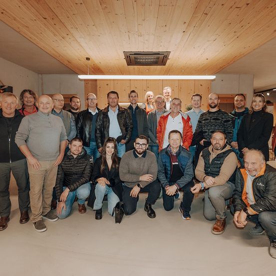 Eine Gruppe von Menschen posiert für ein Foto in einem Raum mit Holzdecken und Beleuchtung. Einige stehen, andere sitzen und sie sind lässig gekleidet.