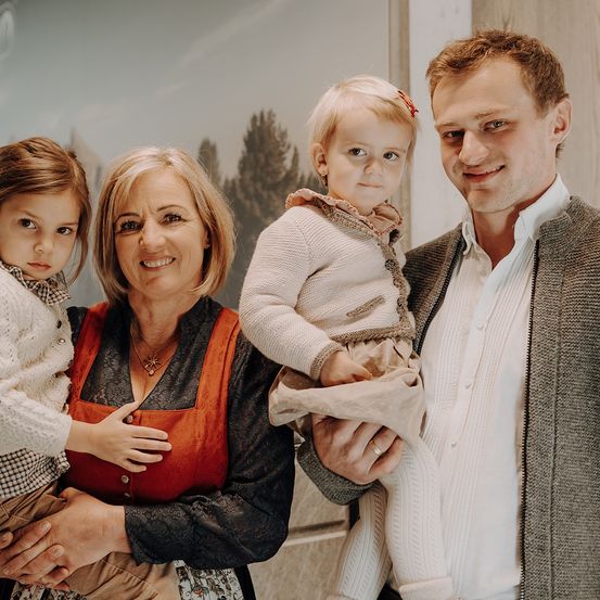 Eine vierköpfige Familie posiert für ein Foto. Eine Frau mit einer Halskette hält ein Kleinkind, während ein Mann ein junges Mädchen hält. Sie stehen nebeneinander und lächeln.