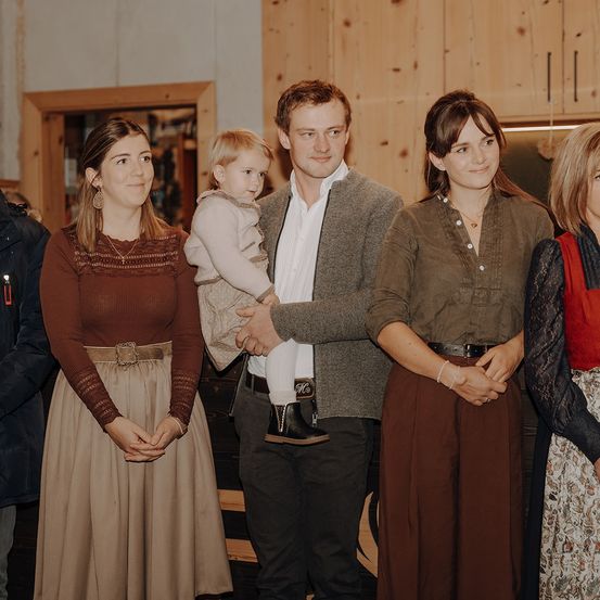 Eine Familie steht in einem Raum mit Holzwänden und lächelt. Ein Mann hält ein Kind, während zwei Frauen auf beiden Seiten stehen.