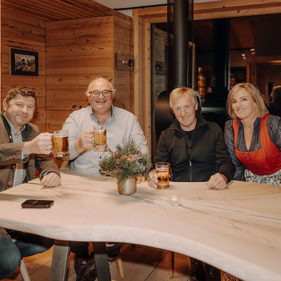 Vier Personen sitzen um einen Holztisch mit Biergläsern herum. Sie scheinen für ein Foto zu posieren. Der Raum hat Holzwände und einen Kamin.