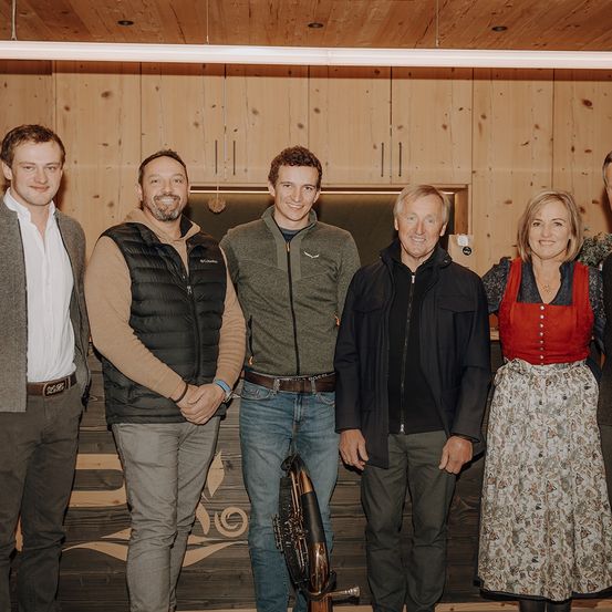 Fünf Personen stehen in einem Raum und lächeln für ein Foto. Eine Trompete befindet sich zwischen zwei Männern. Der Raum hat Holzwände und eine Holztheke.