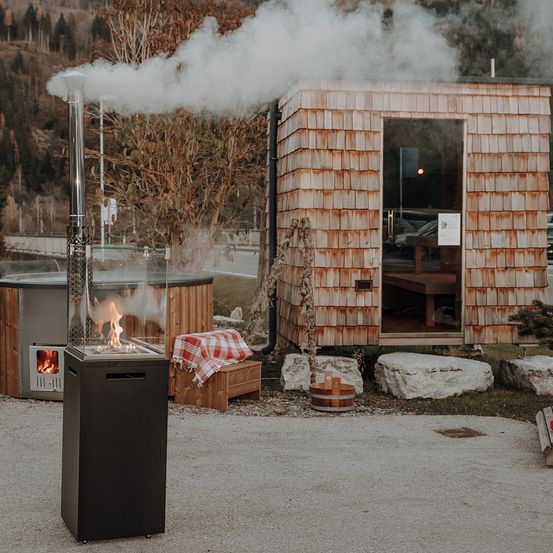 Ein kleines Holzgebäude mit einer Glastür und offenem Eingang gibt Rauch ab. Ein Außenkamin mit Feuer davor. Eine Holzbank mit gemustertem Tuch daneben. Ein Whirlpool hinter dem Gebäude.