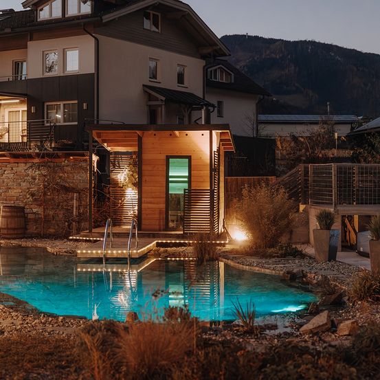 Ein modernes Haus mit einem Pool und einer Holzstruktur mit Lichtern, vor einer Bergkulisse. Das Haus hat Ziegelwände und mehrere Fenster.