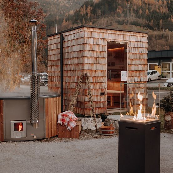 Eine Holzsauna mit Kamin steht im Freien. Ein Feuerkorb ist in der Nähe. Autos sind dahinter geparkt. Bäume und Berge sind in der Ferne.