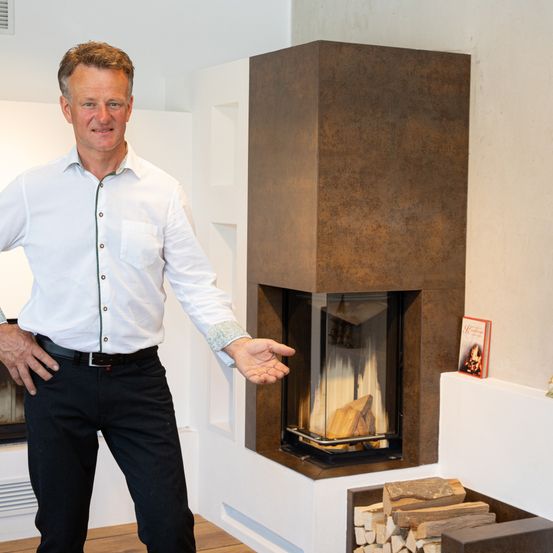 Ein Mann steht neben einem modernen Kamin, in dem ein Feuer brennt, und zeigt darauf. Er trägt ein weißes Hemd und schwarze Hose.