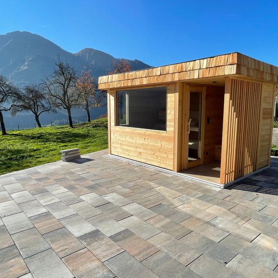 Eine Holzsauna steht draußen auf einem gefliesten Boden, umgeben von Gras und Bäumen mit Bergen in der Ferne.