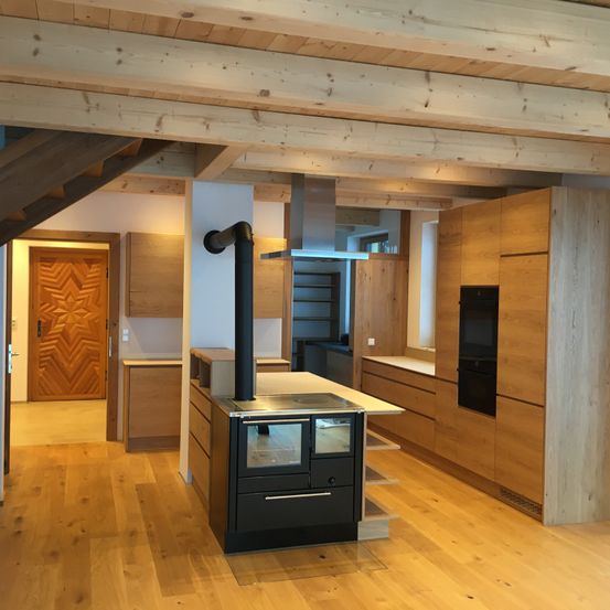Eine moderne Küche mit Holzboden, freiliegenden Holzbalken, einem großen schwarzen Ofen und einer Holztür. Die Küche verfügt über eine zentrale Insel und Schränke.
