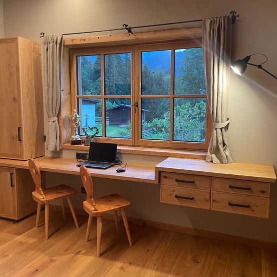 Ein Arbeitsbereich mit Laptop, Trophäe und herzförmigem Objekt auf einem Schreibtisch unter einem Fenster mit Vorhängen. Holzmöbel und eine Lampe.