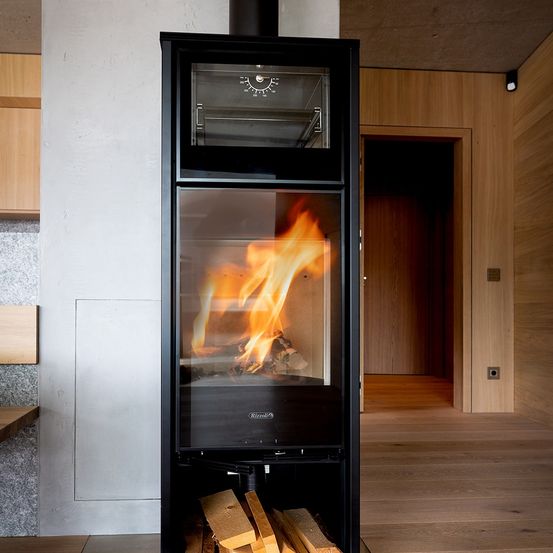 Ein moderner schwarzer Holzofen mit Glastür, die ein Feuer zeigt, steht in einem Raum mit Holzböden und Wänden.