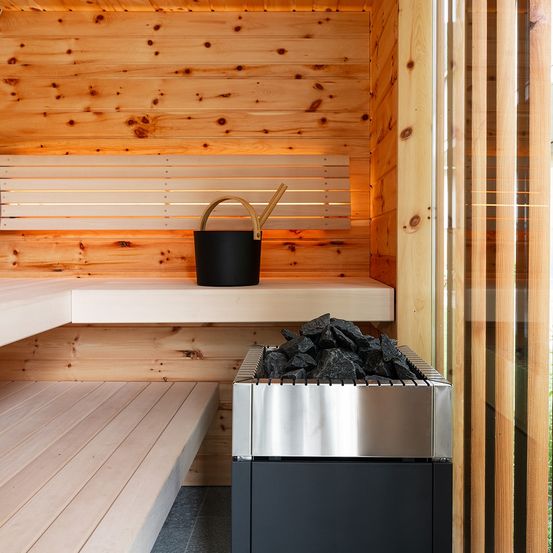 Eine hölzerne Sauna mit einer Bank, einem schwarzen Eimer mit einem Schöpflöffel und einem silber-schwarzen Ofen mit heißen Kohlen. Holzwände und ein Glasfenster sind ebenfalls sichtbar.