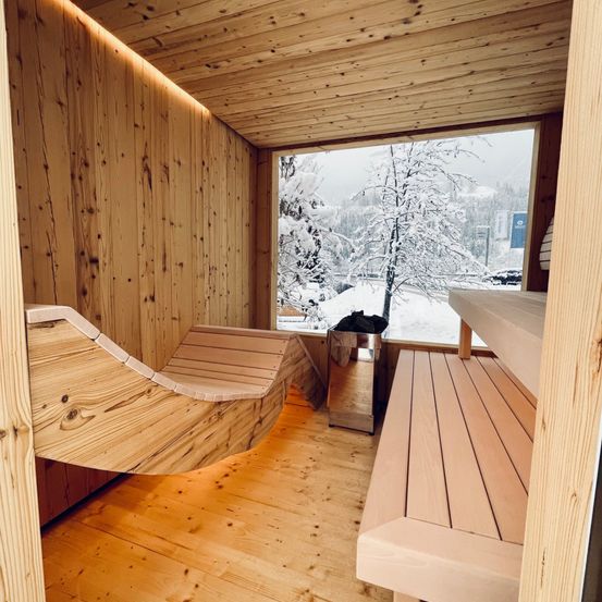 Eine Holzsauna mit einer geschwungenen Bank, einem Mülleimer und einem großen Fenster mit Blick auf eine verschneite Landschaft.