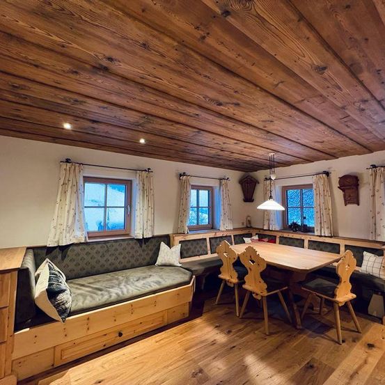 Ein gemütlicher Essbereich mit einem holztischen und Stühlen. Ein Sofa mit Kissen befindet sich unter den Fenstern mit Vorhängen. Holzbalken säumen die Decke und Wände.