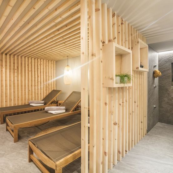 Ein holzverkleideter Spa-Raum mit Liegen und Regalen. Der Raum wird von Sonnenlicht erhellt.