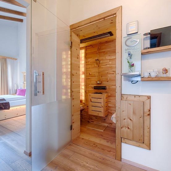 Innenansicht eines modernen Hotelzimmers mit Holzboden, Glastür zur Sauna und Holzregalen mit Tassen und Gegenständen.