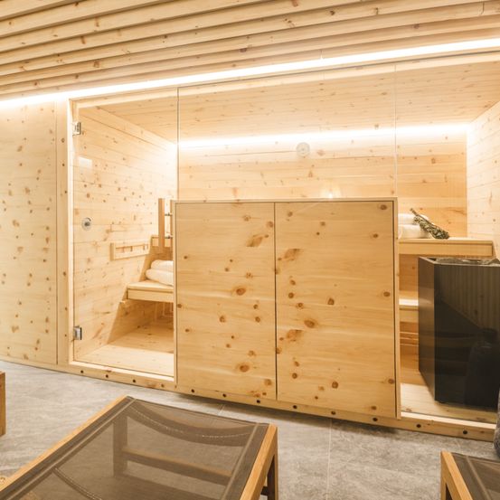 Ein holzverkleideter Saunaraum mit einer Tür, die zu einem Sitzbereich führt, Holzregalen, einer Glastür und einer Holzdecke.