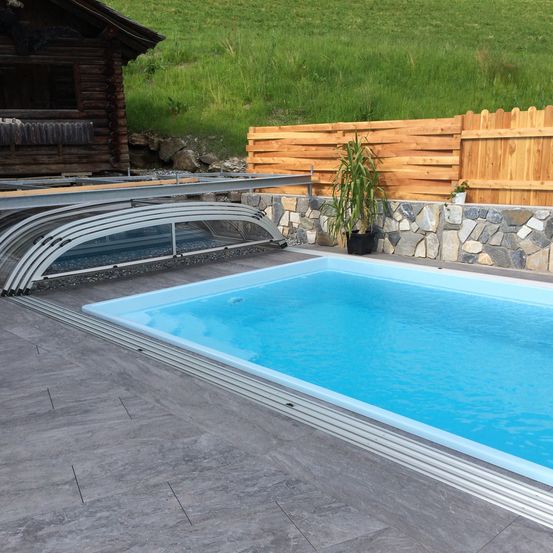 Ein Außenpool mit Glasabdeckung und Holzzaun im Garten mit einem Blockhaus und grünem Rasen.