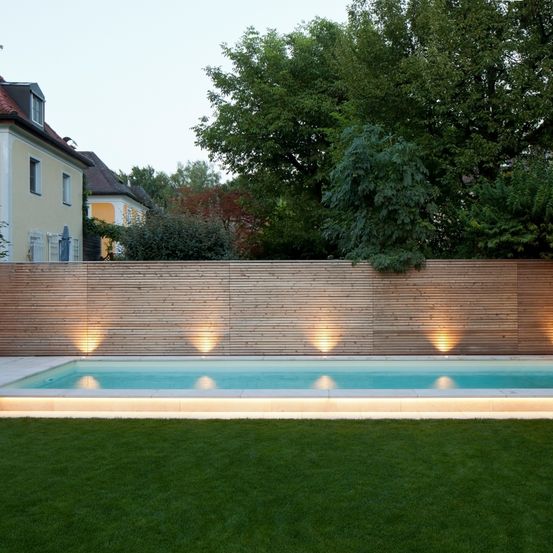 Ein Garten mit einem Holzzaun und einem Pool, umgeben von einem Rasen. Im Hintergrund befinden sich Bäume und Büsche. Der Pool hat Lichter an seinen Seiten.