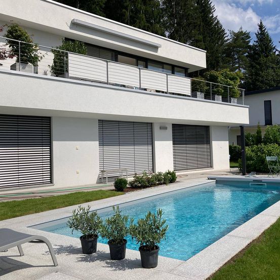 Modernes weißes Haus mit Swimmingpool, Balkon mit Geländer und Topfpflanzen. Zwei Liegestühle links und ein Stuhl rechts.