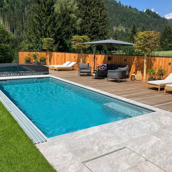 Ein moderner, rechteckiger Swimmingpool mit einer Holzterrasse, umgeben von einem Holzzaun. Zwei Liegestühle und eine Couch mit Kissen sind um einen Tisch herum aufgestellt, und ein Sonnenschirm ist in der Nähe positioniert.