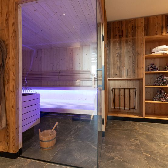 Ein holzverkleideter Saunaraum mit lila Beleuchtung, einer Glastür, einem Holzeimer und Regalen mit Gegenständen.