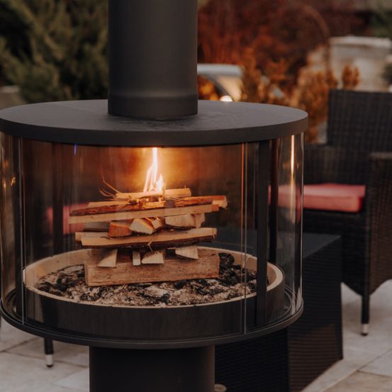 Ein moderner Außenkamin mit brennendem Feuer darin. Er steht auf einer Terrasse mit einem Korbstuhl in der Nähe.