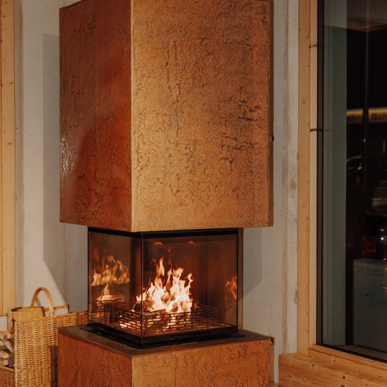Ein moderner Kamin mit einer braunen strukturierten Außenseite, einer Glastür und einem brennenden Feuer im Inneren. Er steht in einem Raum mit Holzwänden und einem Fenster.