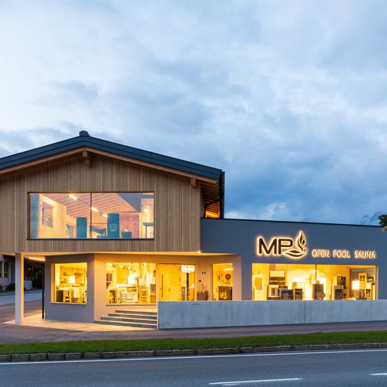 Ein modernes Gebäude mit Holzfassade und Glasfenstern ist nachts beleuchtet. Es trägt ein Schild mit der Aufschrift 'MP Open Pool Sauna'. Das Erdgeschoss hat eine Glaswand und eine Tür.