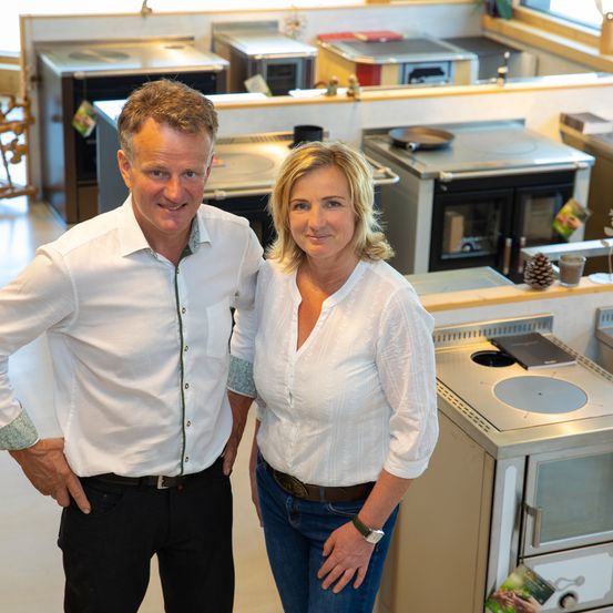 Ein Mann und eine Frau stehen lächelnd für die Kamera in einem Küchenbereich. Sie sind von verschiedenen Küchengeräten und Dekorationen umgeben.