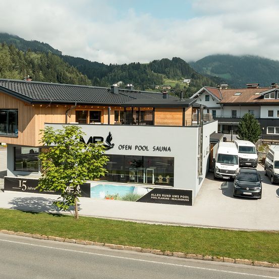 Ein modernes Gebäude mit schwarzem Dach, einer weißen Wand mit Pool-Logo und einem Baum davor. Vor dem Gebäude sind Fahrzeuge geparkt.