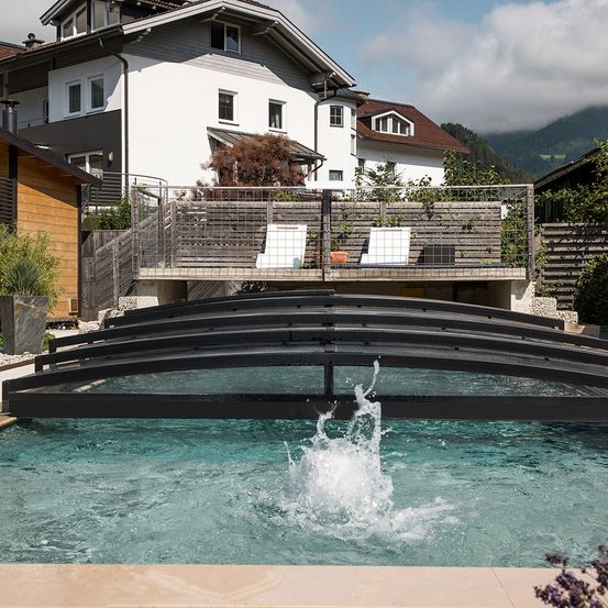 Ein moderner Swimmingpool mit einer schwarzen Abdeckung vor einem Haus. Es gibt einen Balkon mit einem Metallzaun und Blick auf die Berge.