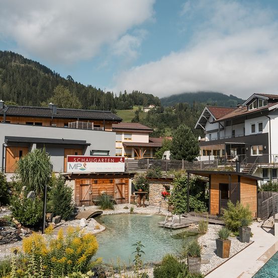 Ein ruhiger Bergurlaub mit Schaugarten, einem Pool, Holzhäusern und üppiger Grünfläche vor einer Kulisse von Bergen und einem bewölkten Himmel.