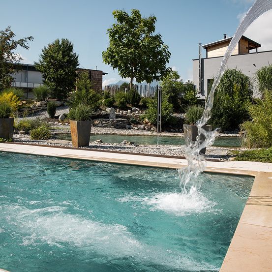 Ein Swimmingpool mit Wasserfluss. Der Bereich ist von Bäumen, Pflanzen und Felsen umgeben. Hinter dem Pool steht ein Haus mit Schornstein.