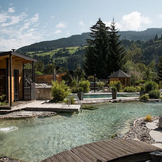 Ein ruhiger Außenbereich mit klarem Wasser, umgeben von Grün und Holzstrukturen. Im Hintergrund befinden sich Berge und ein blauer Himmel.