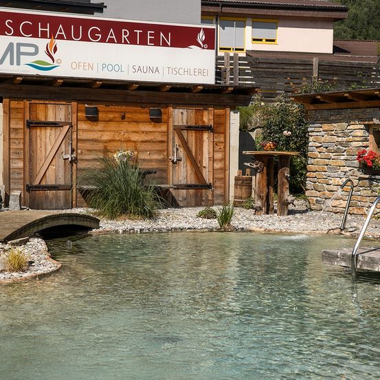 Ein Freibereich mit Schwimmbecken, Holzgebäuden und einem Schild mit der Aufschrift Schaugarten. Das Becken hat eine Brücke auf der linken Seite.