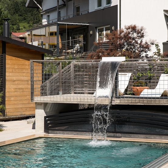 Ein moderner Pool mit einem Wasserfall, der hineinfließt, umgeben von einem Metallzaun, und einem Gebäude mit Balkon und Treppen dahinter.