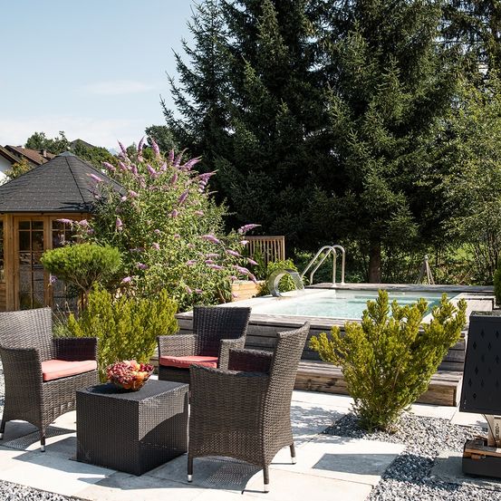Eine Terrasse mit Tisch, Korbstühlen und einem Swimmingpool mit Holzdeck in einem Garten.