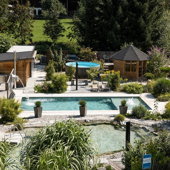 Luftaufnahme eines ruhigen Gartens mit einem großen Swimmingpool, einem kleinen Holzhaus und verschiedenen Topfpflanzen. Grünes Gras, Bäume und ein blauer Himmel im Hintergrund.