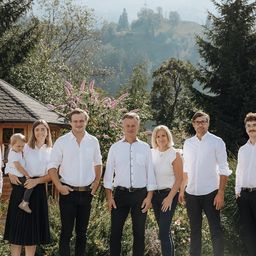 Eine Familie steht in einem Garten mit üppiger Vegetation und Bergen im Hintergrund. Sie sind in passenden weißen Hemden und dunklen Hosen gekleidet.