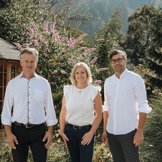 Drei Personen, zwei Männer und eine Frau, stehen zusammen in einem Garten mit rosa Blumen und Grün, lächeln für ein Foto.