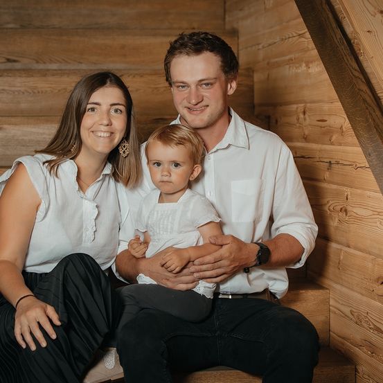 Eine lächelnde Familie aus drei Personen, bestehend aus einer Frau, einem Mann und einem kleinen Kind, sitzen auf einer Holztreppe in einem gemütlichen Zimmer.