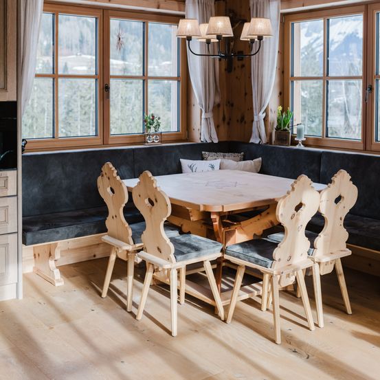 Ein Holztisch mit vier Stühlen steht unter einer Kronleuchter in einem Essbereich. Es gibt eine Bank an der Wand und Fenster lassen natürliches Licht herein. Draußen gibt es Bäume und Berge.