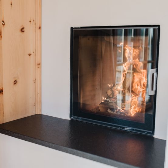 Ein moderner Kamin mit brennendem Feuer darin. Der Kamin ist an einer weißen Wand montiert und der Boden ist schwarz.