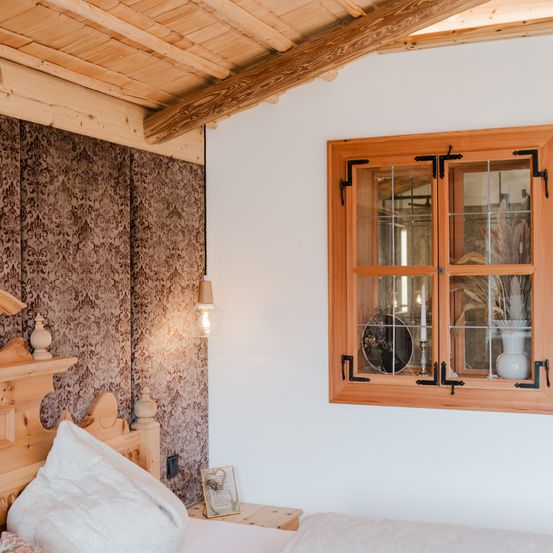 Ein Schlafzimmer mit einem Holzbett, Kissen und einem Nachttisch. Eine gemusterte Tapete und ein Holzfensterrahmen sind sichtbar. Eine Lampe ist an der Wand montiert und ein Bilderrahmen steht auf dem Nachttisch.