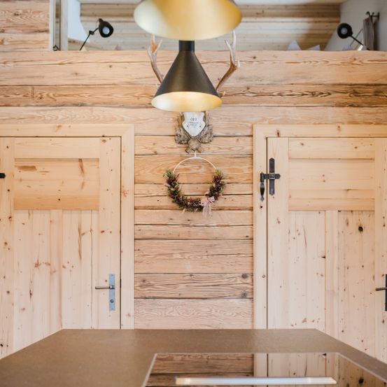 Innenansicht einer Holzhütte mit Holzwänden, einer Lampe, zwei Türen und einem Kranz an der Wand.