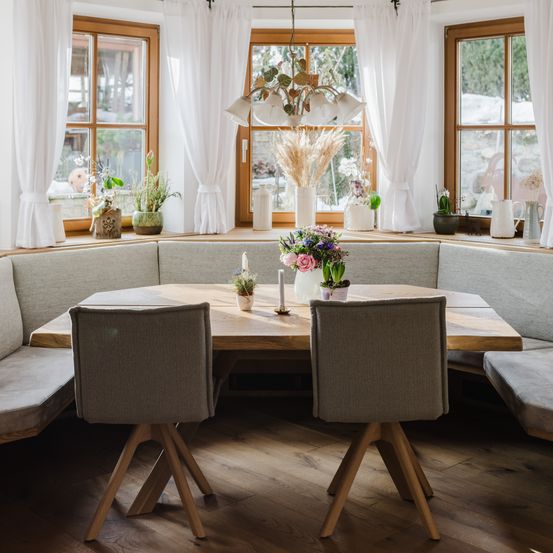 Ein gemütlicher Essbereich mit einem Holztisch und Stühlen, einer Bank und einem Fenster mit weißen Vorhängen. Der Tisch hat Blumen, eine Kerze und Pflanzen darauf.