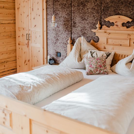 Ein Doppelbett mit hölzerne Kopfteil und gemusterte Tapete. Zwei Kissen, eines mit Hirschgeweih, liegen auf dem Bett. Ein Schrank und eine Lampe befinden sich links.