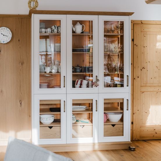 Eine Küchenvitrine mit Glastüren, Holzschubladen und Regalen, die verschiedene Küchenutensilien wie Schüsseln, Tassen, Teller und Teekannen präsentiert. Die Umgebung ist warm mit Holzwänden und -böden.