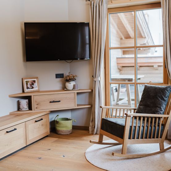 Ein gemütliches Zimmer mit Holzmöbeln, einem Flachbildfernseher an der Wand, einem Schaukelstuhl und einem Glasfenster mit Blick nach draußen.